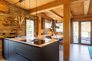 Una cocina moderna con una isla central, pared de piedra, vigas de madera e iluminación colgante, y vistas al exterior.
