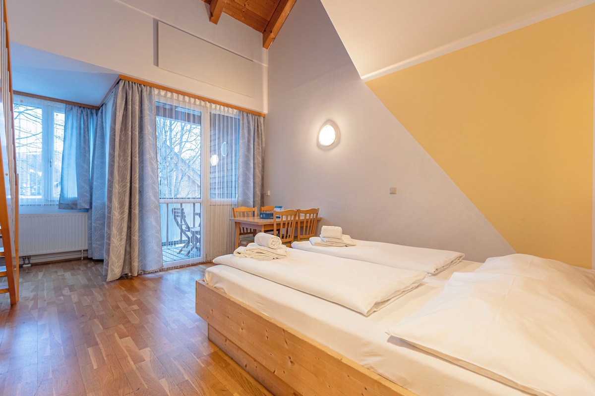 A bedroom with a large bed, a wooden floor, a table with chairs, and a view of the outside through a window.