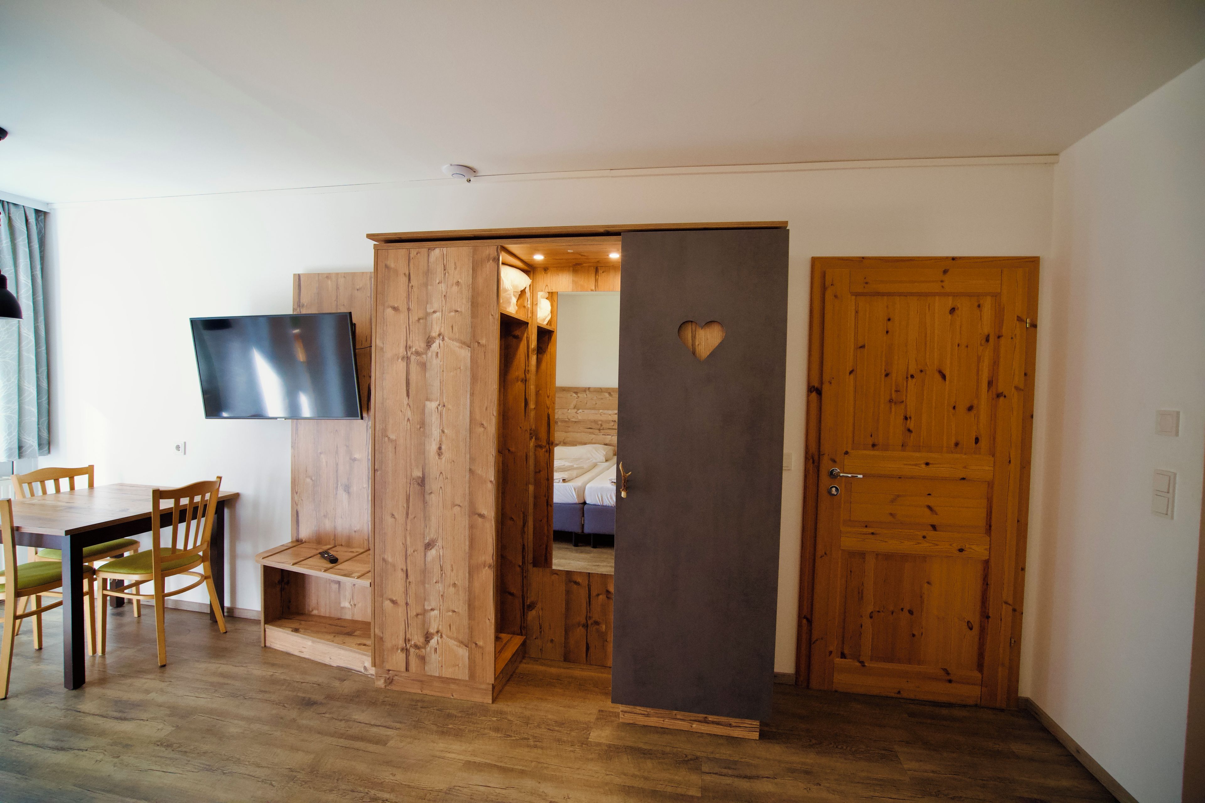A room with a wooden floor, a TV mounted on the wall, a wooden door with a heart cutout, and a bed with a headboard.