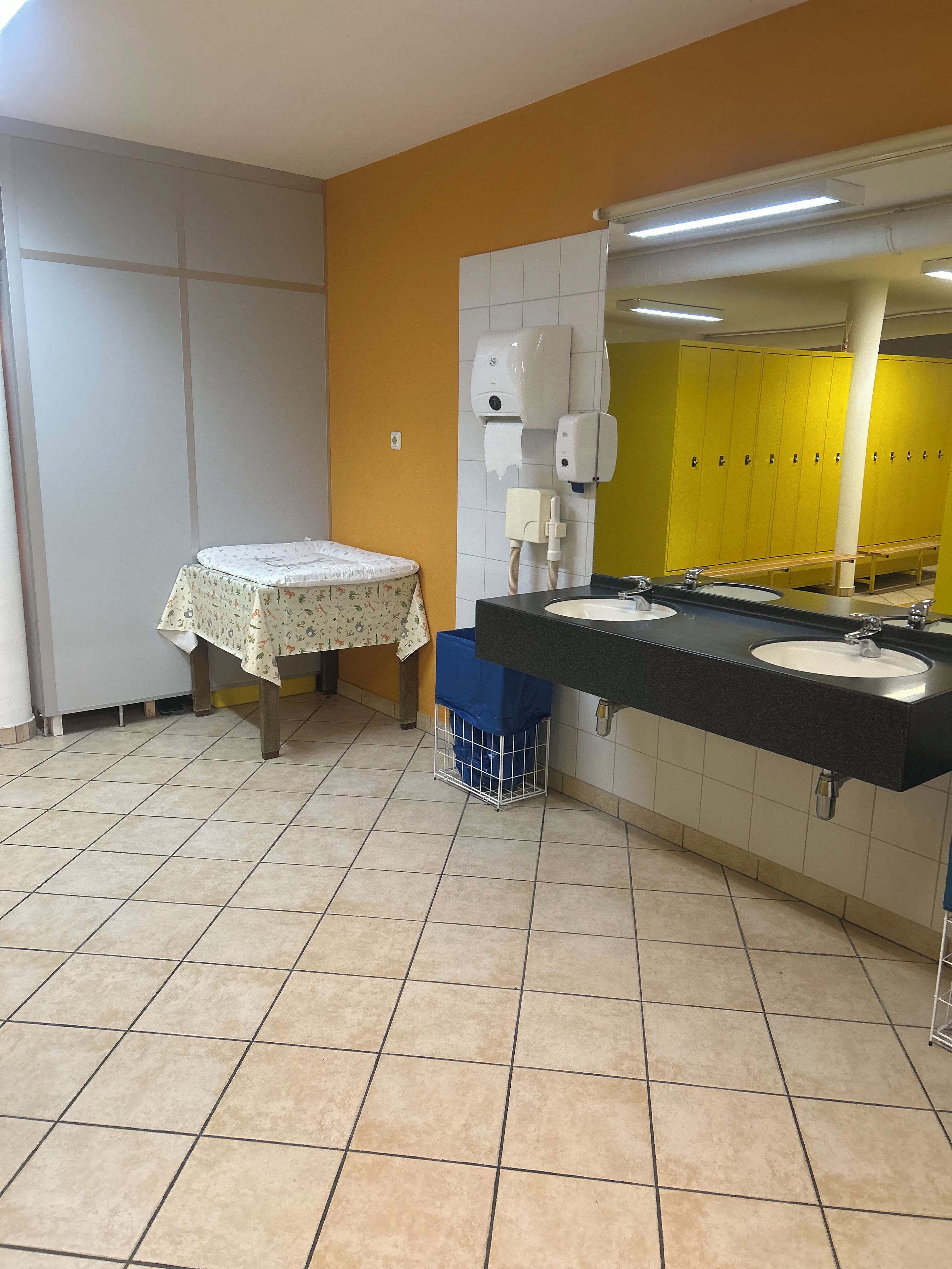 Des toilettes avec deux lavabos, une table recouverte d’une nappe à motifs floraux et une rangée de casiers jaunes.