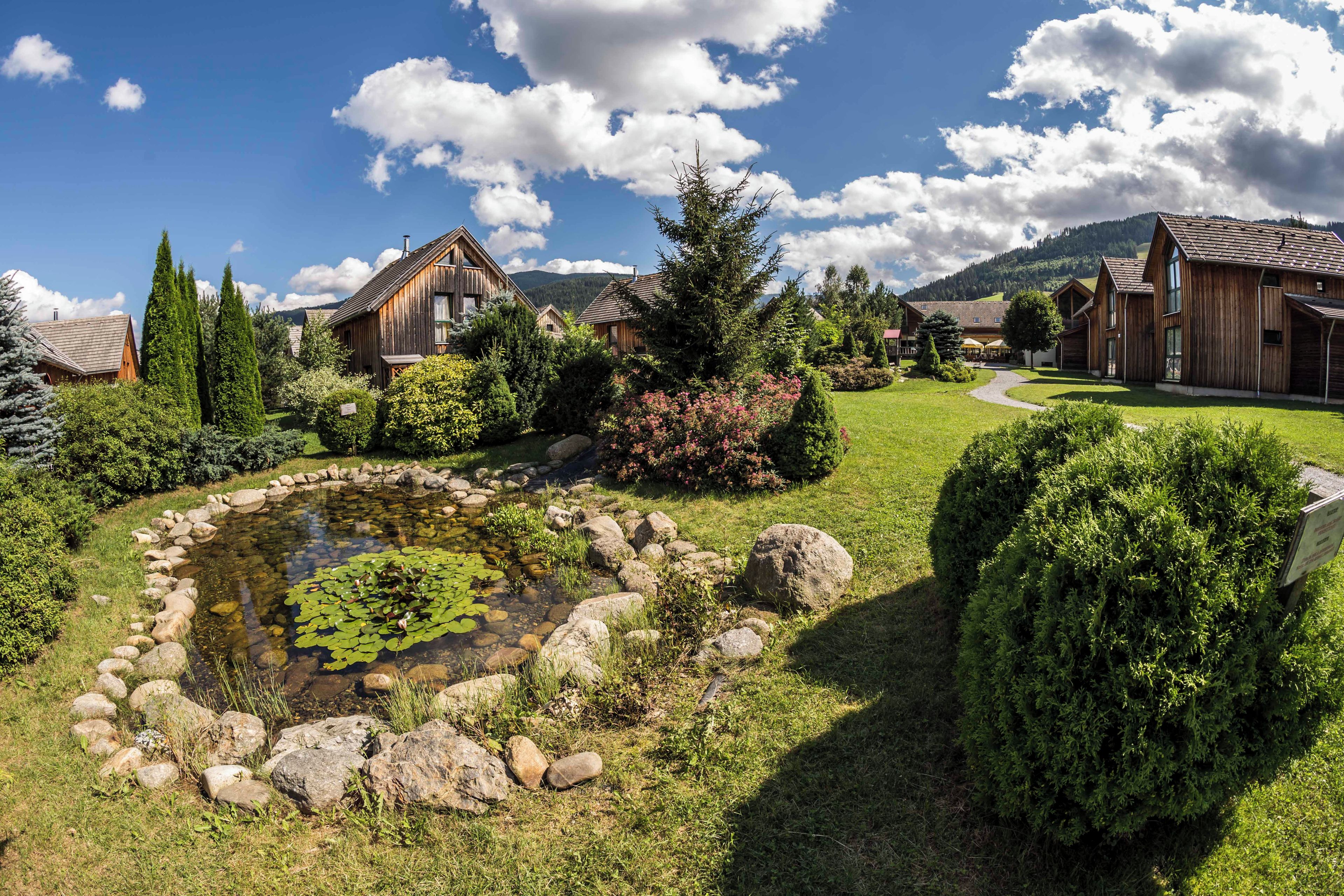 Un jardín tranquilo con un estanque, rodeado de casas de madera y abundante vegetación.