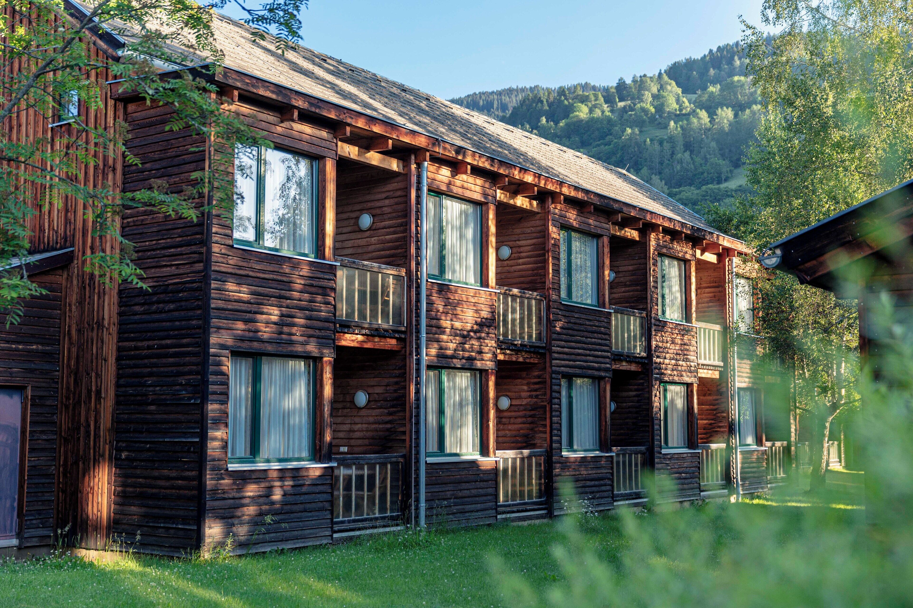 A multi-story log cabin with balconies, surrounded by greenery and mountains.