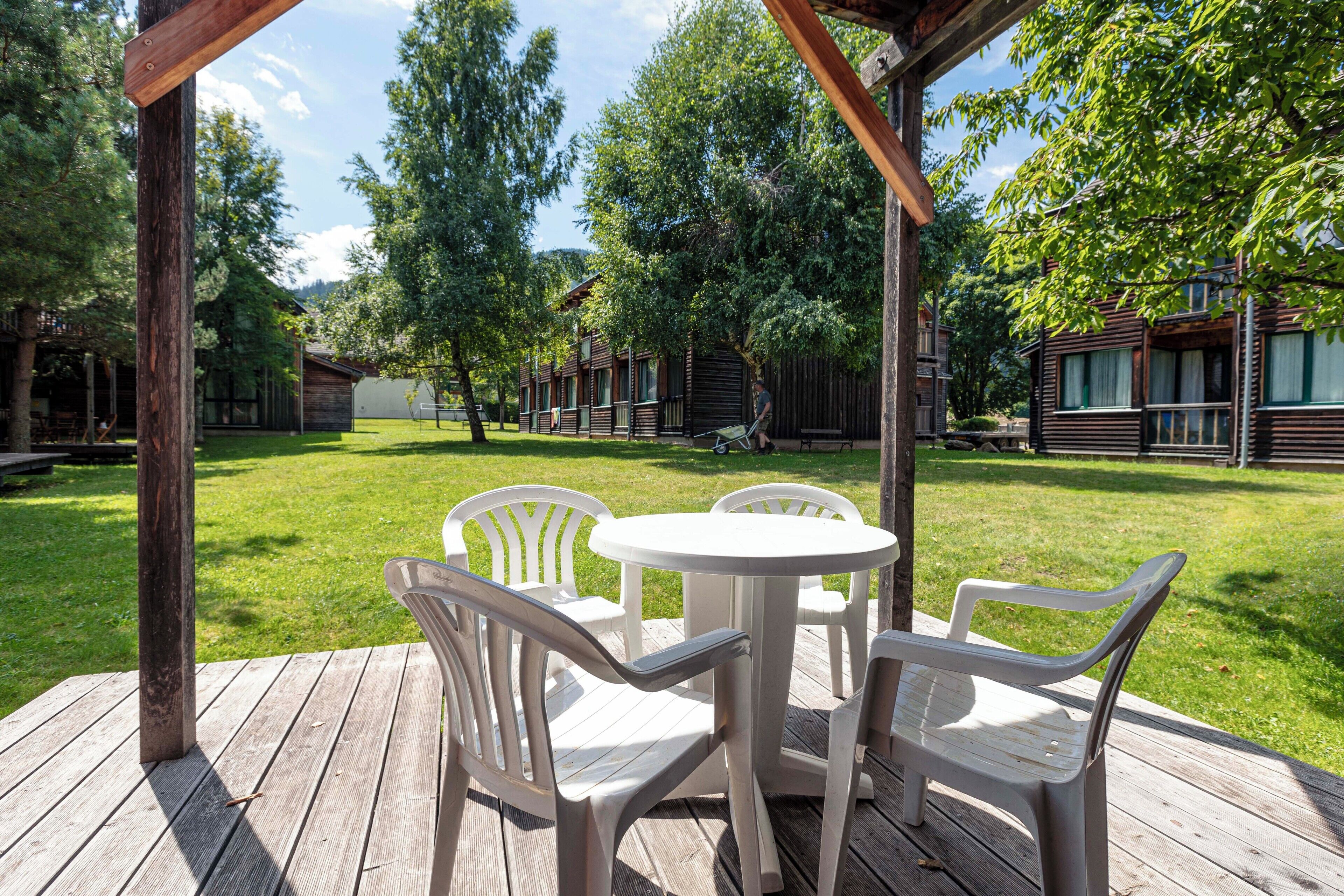 A wooden deck with a round table and four chairs, surrounded by a grassy area with trees and cabins in the background.