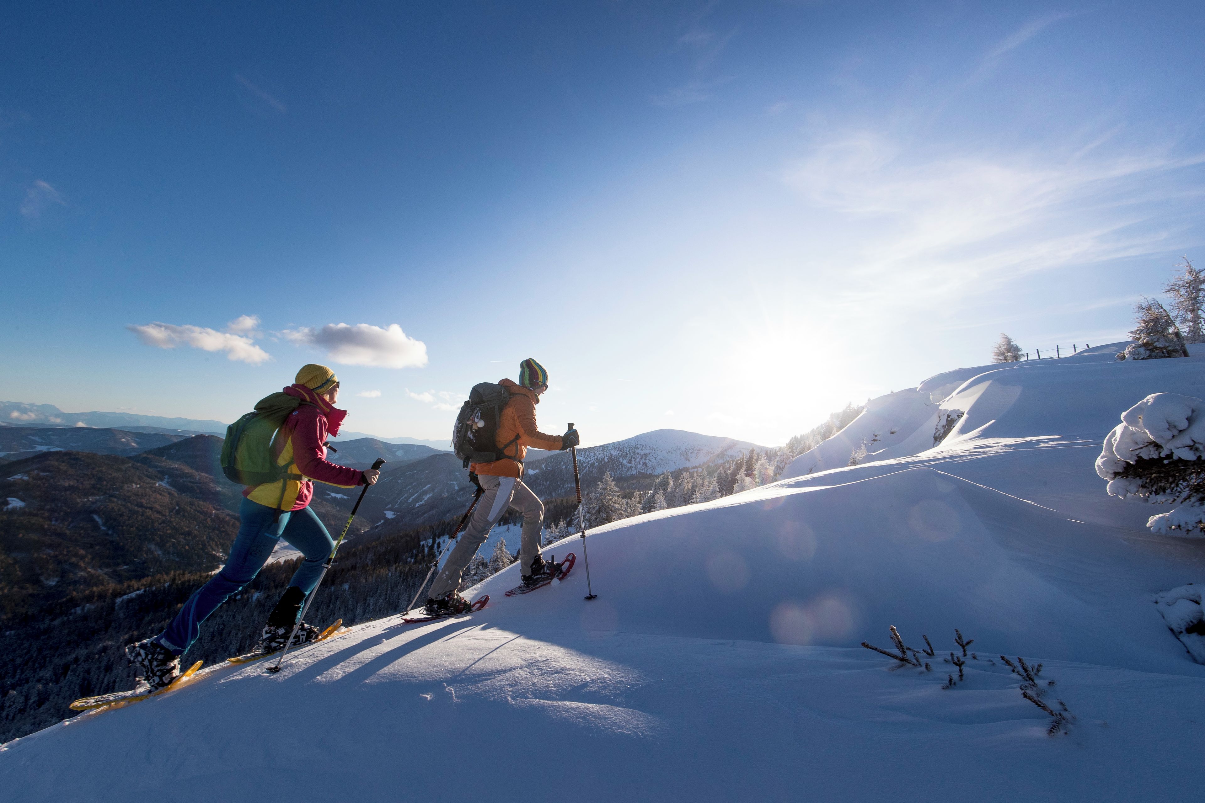 Two people hiking on a snow-covered slope with a clear sky and distant mountains.