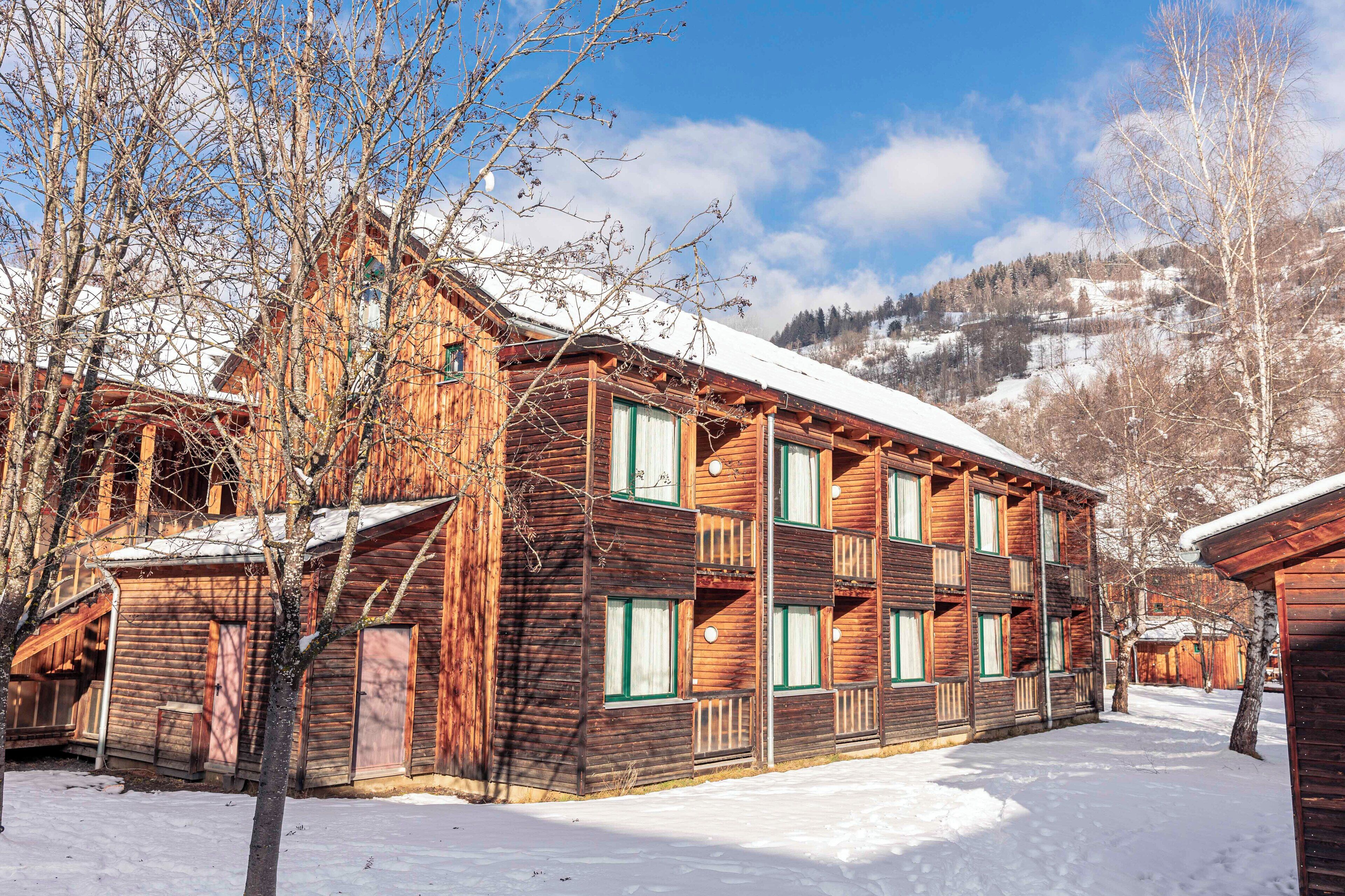 A wooden building with multiple windows and balconies, surrounded by snow-covered ground and bare trees.