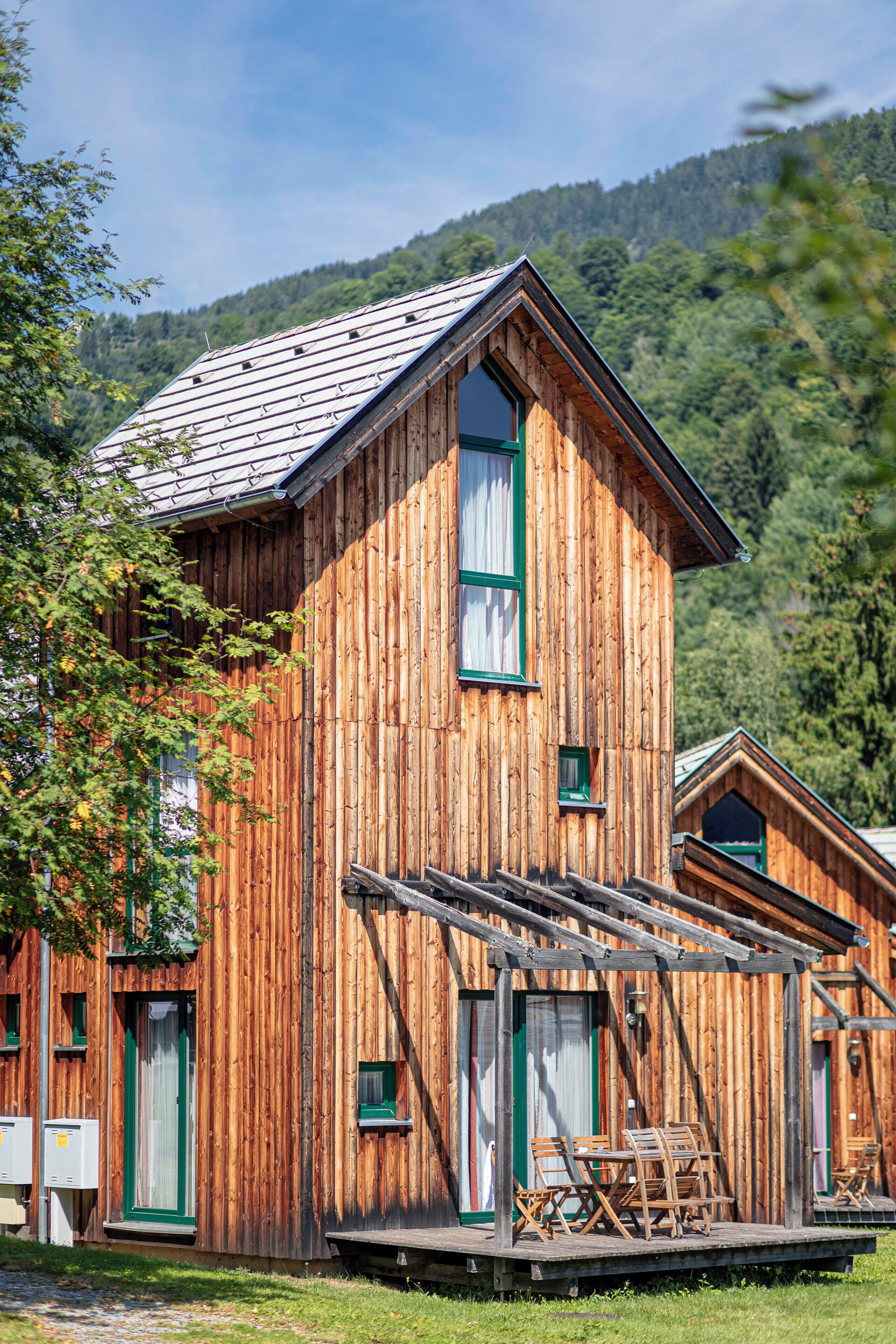 A wooden cabin with a sloped roof, large windows, and a porch with chairs.