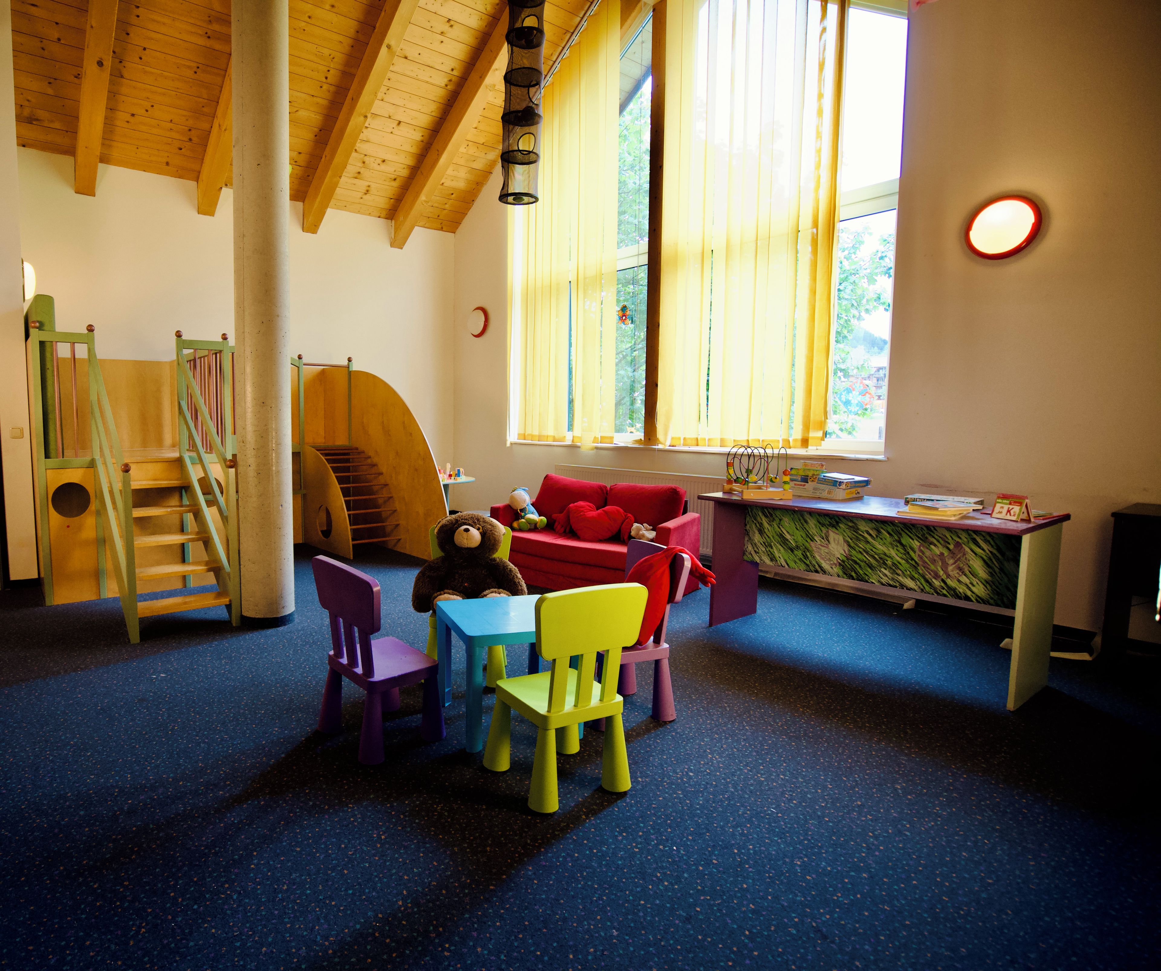 A children's playroom with a wooden staircase, colorful chairs, a table, and a window with curtains.