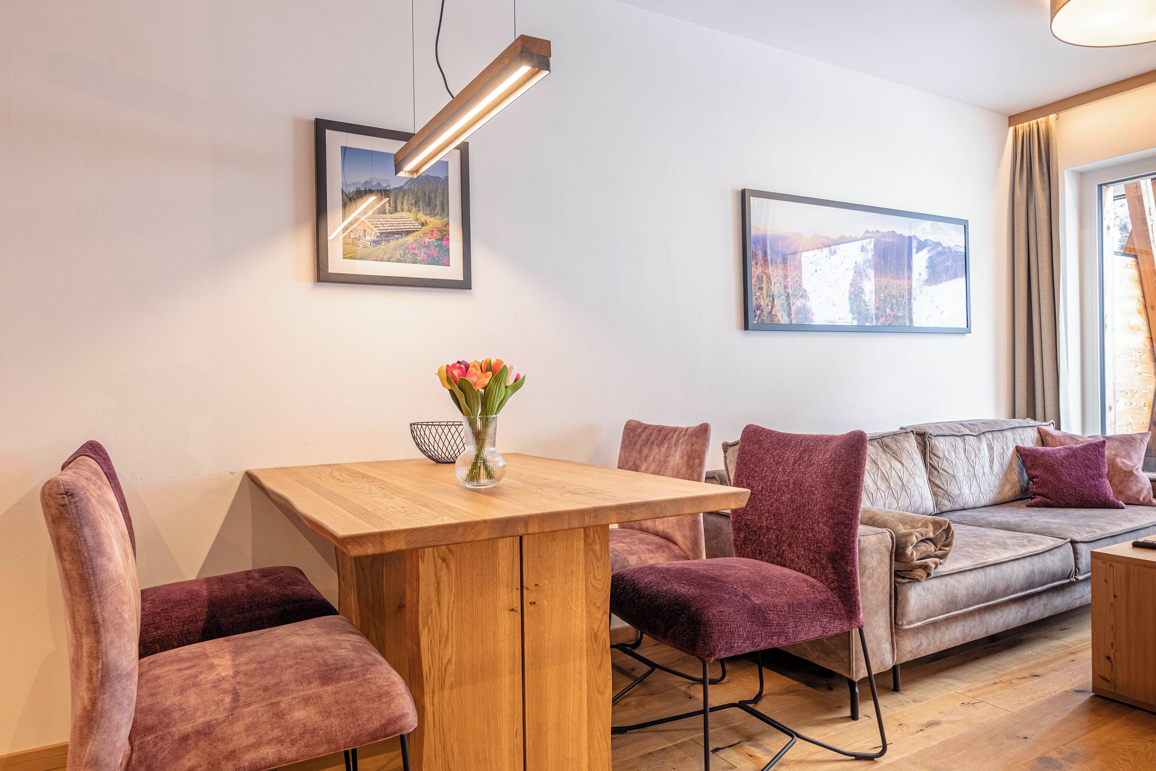 Una sala moderna con una mesa de comedor de madera, sillas tapizadas en color morado, un sofá con cojines y dos cuadros enmarcados en la pared.