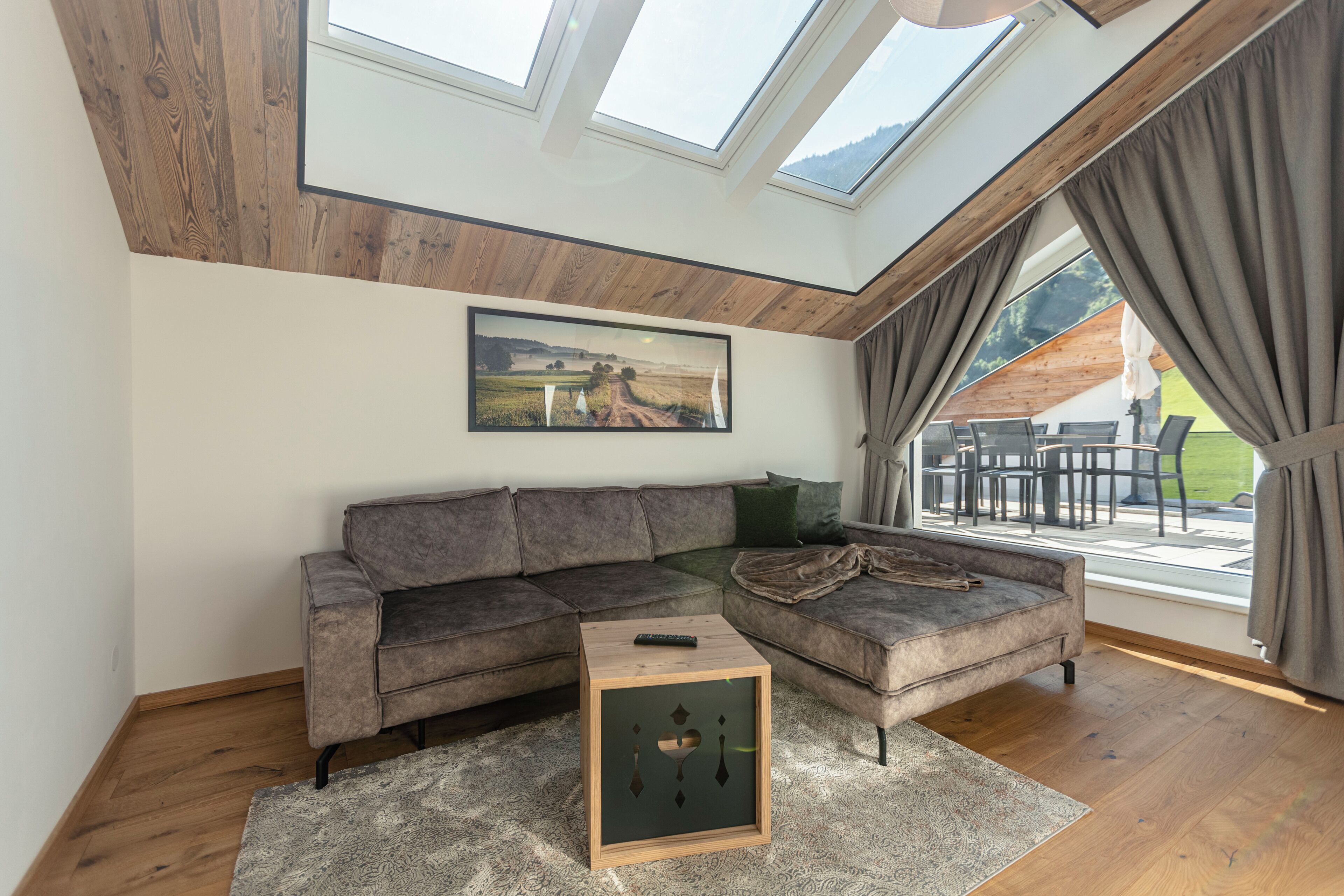 A modern living room with a wooden ceiling, a large window with curtains, a grey sofa, a small wooden side table, and a framed picture on the wall.