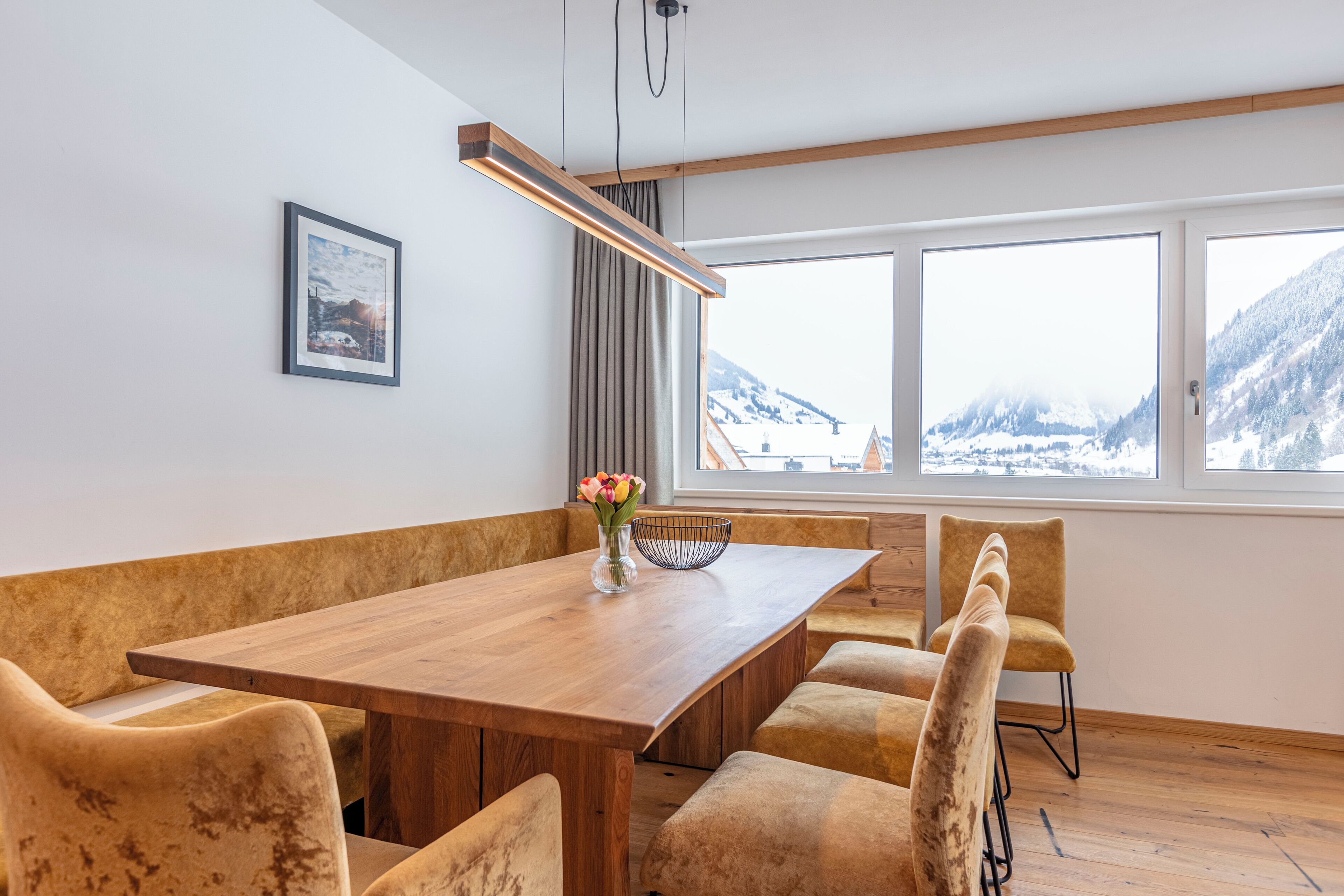 Un comedor con mesa de madera, sillas acolchadas, un jarrón con flores y vistas a montañas nevadas a través de amplias ventanas.
