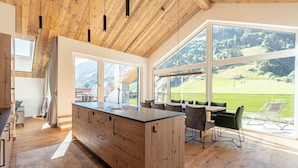 A modern kitchen with a large island, wooden ceiling, and large windows offering a view of a green landscape. - Carpe Solem Rauris by ALPS RESORTS (Rauris)