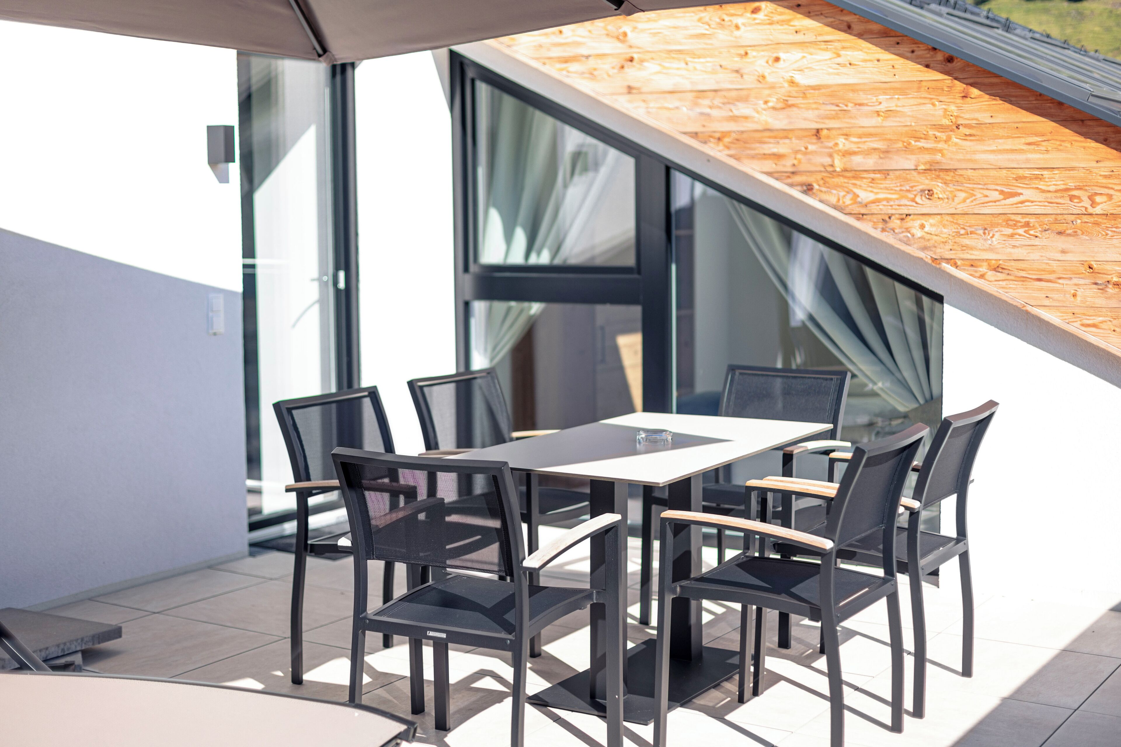 Een buitenterras met een tafel, stoelen, een schuin houten dak en grote ramen.