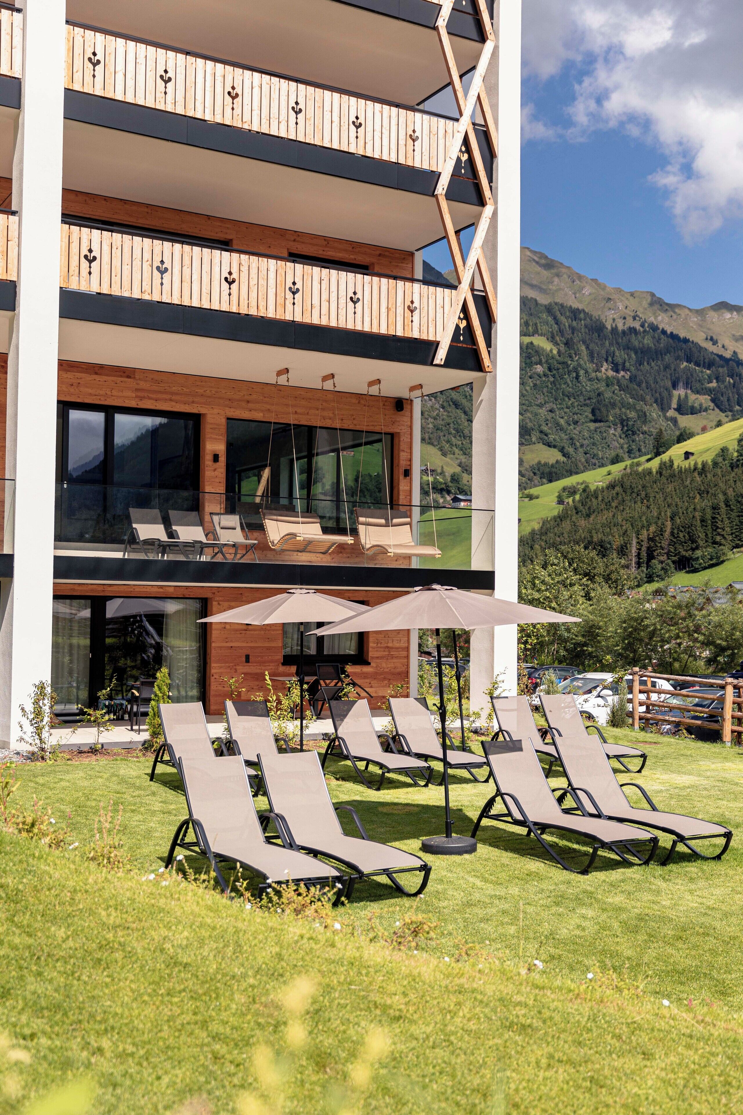 Un edificio moderno con balcón de madera y zona de estar al aire libre con sillones y sombrillas.