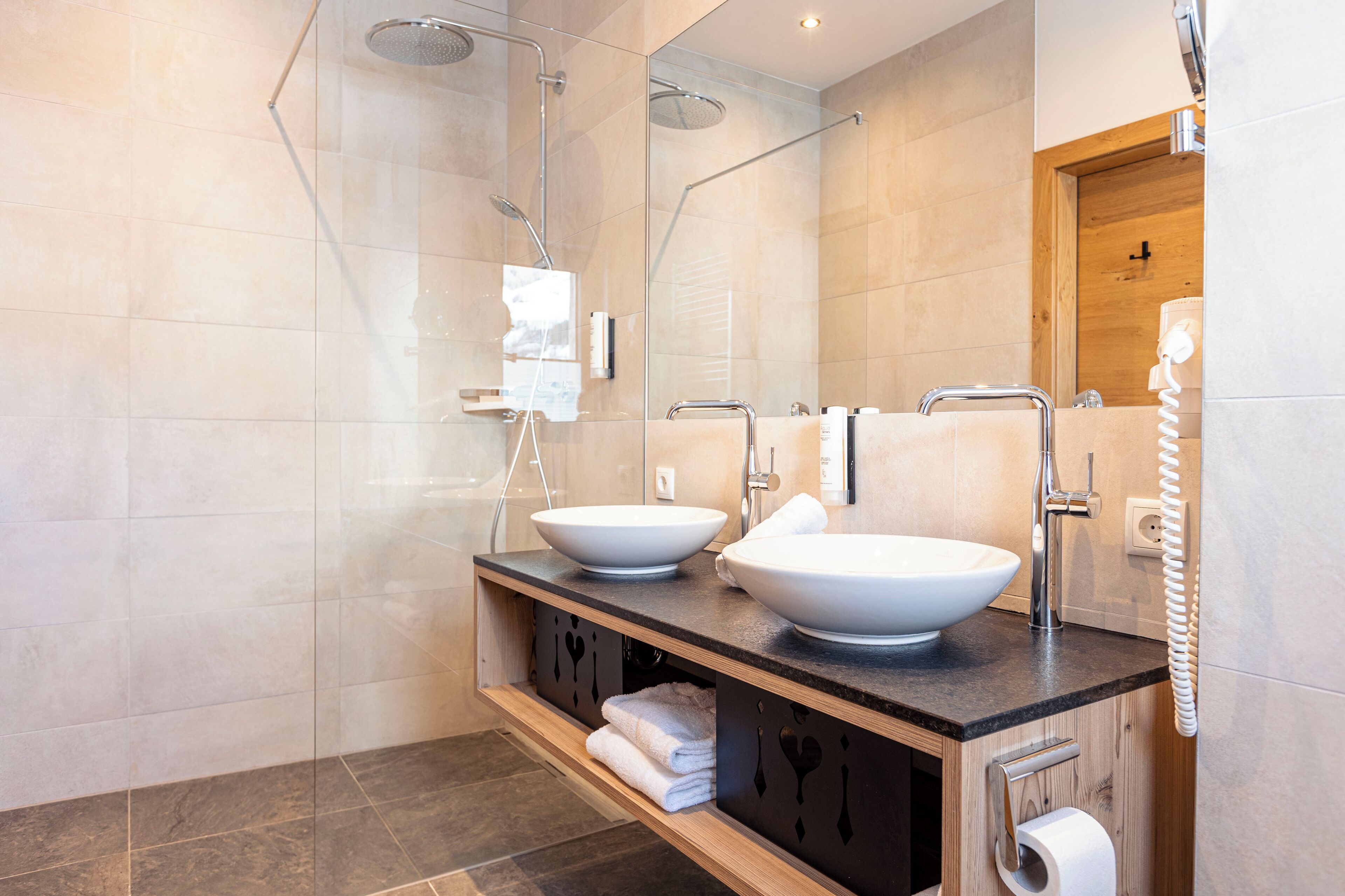 Un baño moderno con dos lavamanos blancos grandes, una ducha con mampara de vidrio y un mueble de tocador de madera con toallas.