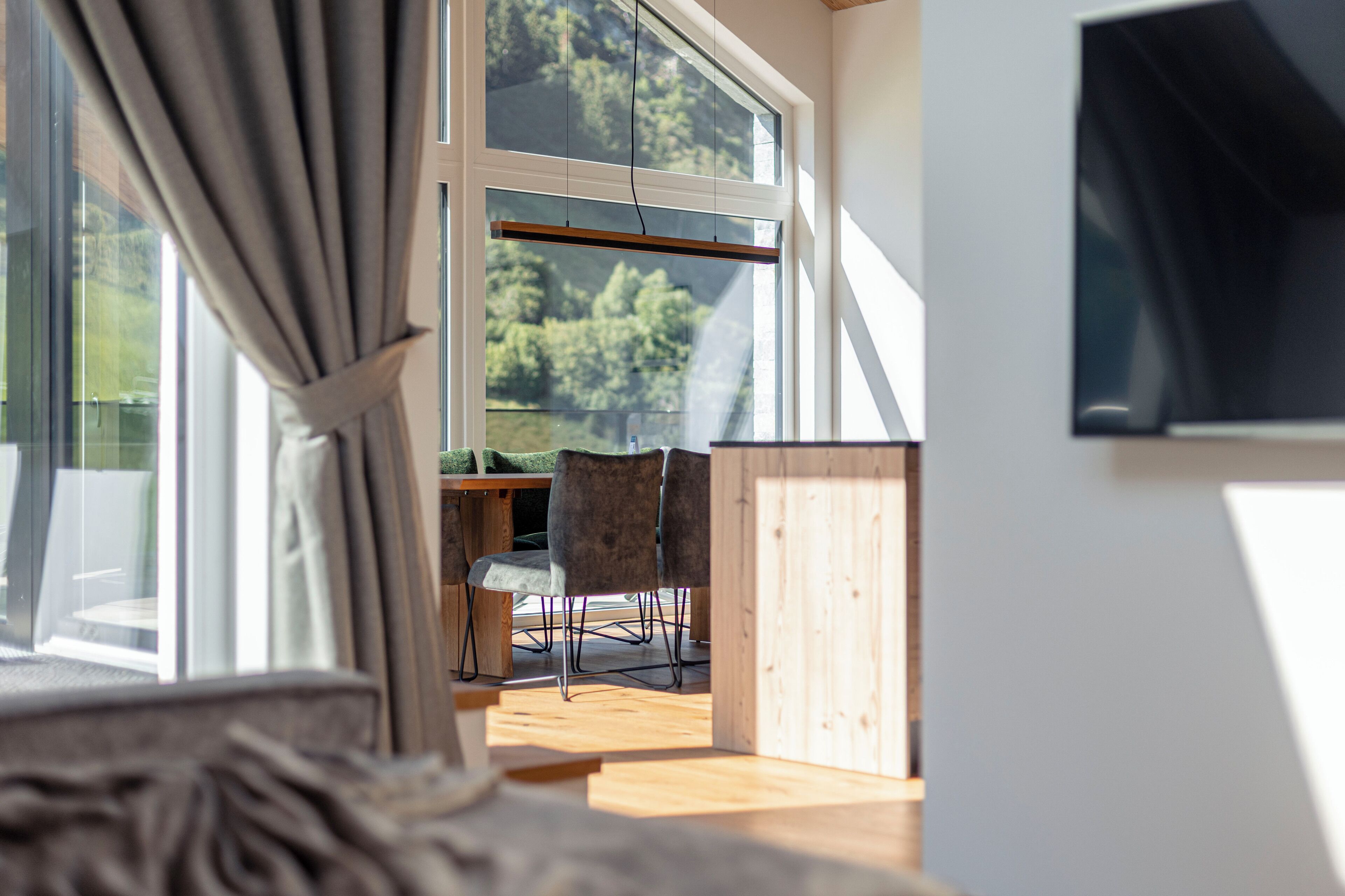 Una habitación con un ventanal grande, piso de madera, un escritorio con sillas y un televisor colocado en la pared.