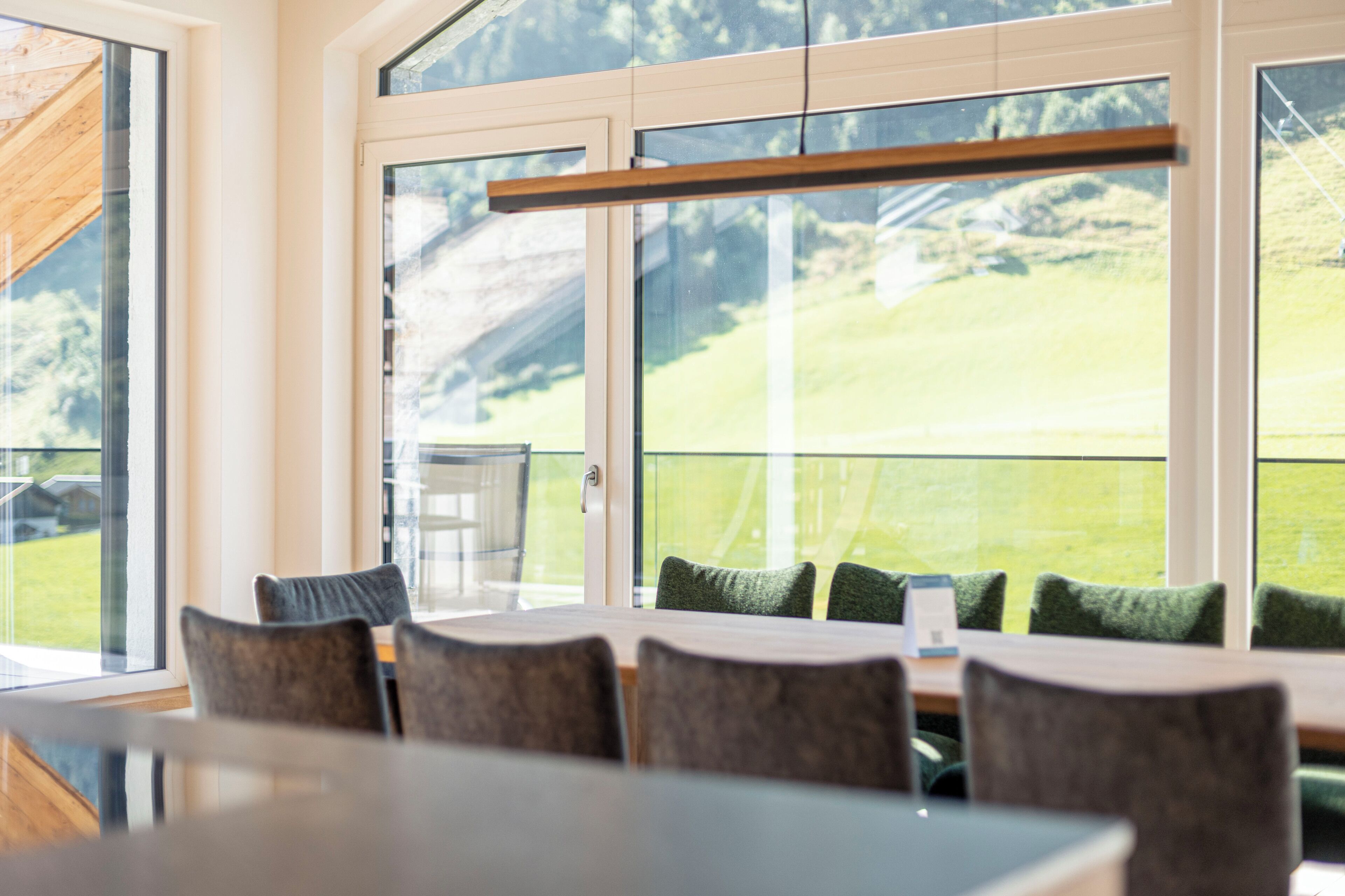 A dining area with a large table and chairs, large windows with a view of a green landscape, and a wooden beam above the table.