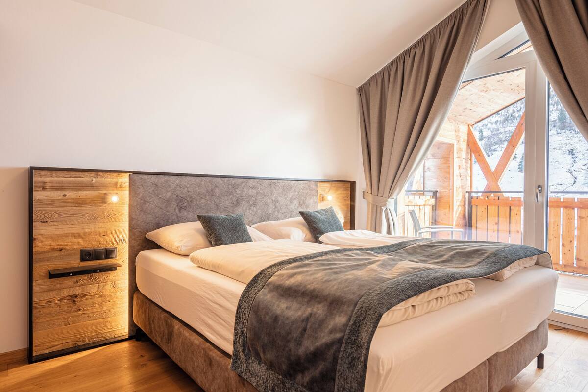 A modern bedroom with a wooden headboard, a large bed with a grey blanket, and a view of a wooden deck and snowy landscape through a glass door.