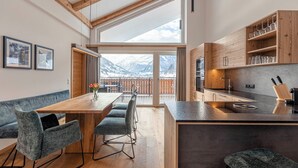 A modern kitchen with wooden cabinets and a large island, a dining area with a wooden table and chairs, and a view of snow-capped mountains through large windows. - Carpe Solem Rauris by ALPS RESORTS (Rauris)