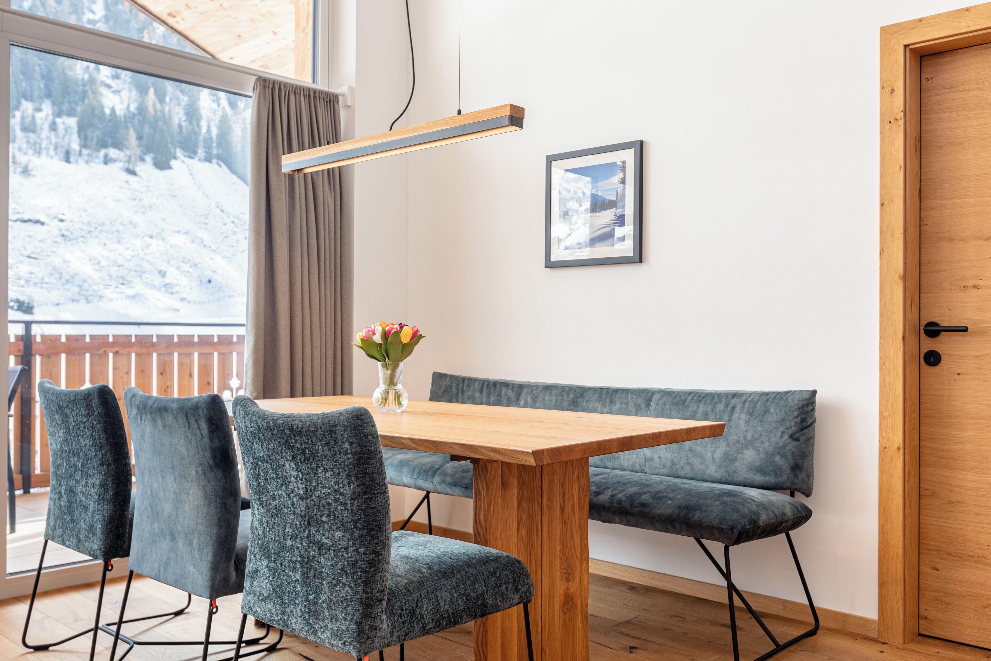 Ein moderner Essbereich mit Holztisch, grau gepolsterten Stühlen, einer Bank, einer Vase mit Blumen, einem Bild an der Wand und Blick auf eine verschneite Landschaft durch ein großes Fenster.