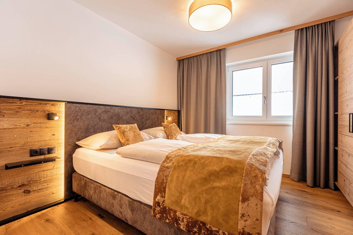 A bedroom with a wooden headboard, a bed with white linens and a brown throw, a bedside table with a lamp, and a window with curtains.