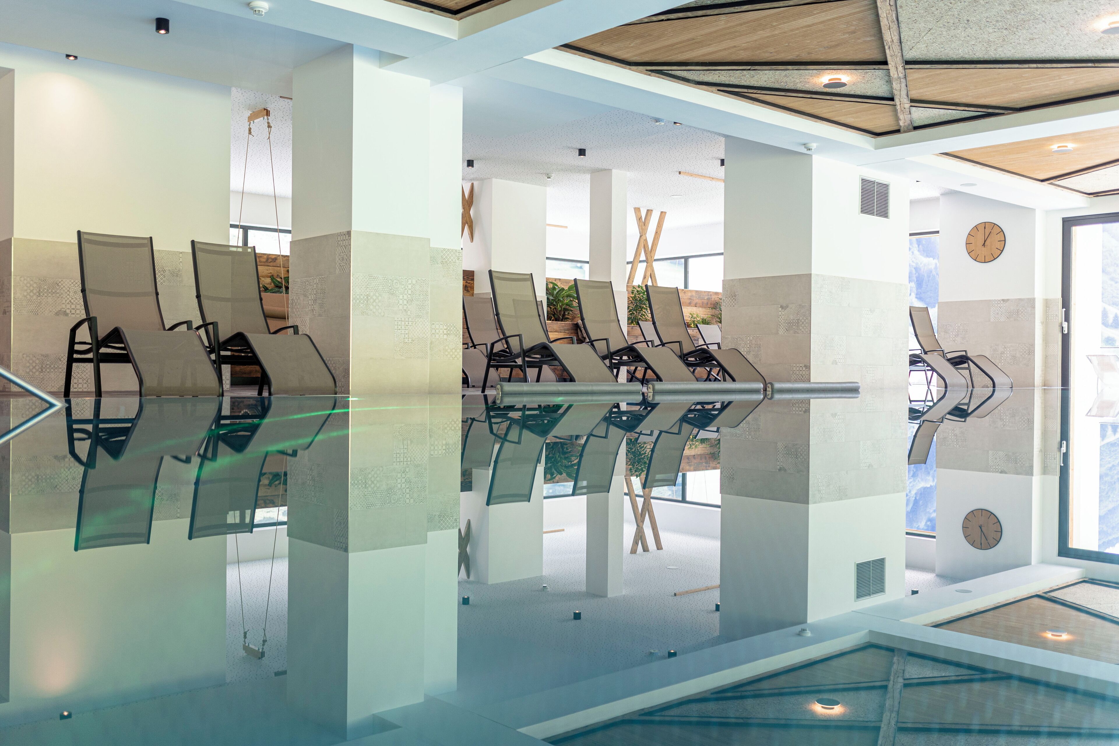 A modern indoor pool area with glass walls and ceiling, featuring multiple lounge chairs and a visible clock on the wall.