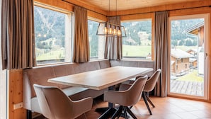 Una cabaña de madera con una gran mesa de comedor y bancos, ventanas amplias con cortinas y vistas a la montaña.