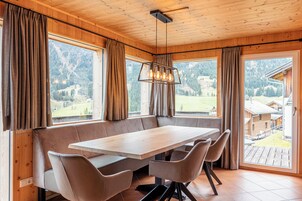 Una cabaña de madera con una gran mesa de comedor y bancos, ventanas amplias con cortinas y vistas a la montaña.