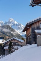 Eine Holzhütte mit schneebedecktem Dach, vor einer Kulisse aus schneebedeckten Bergen und blauem Himmel.