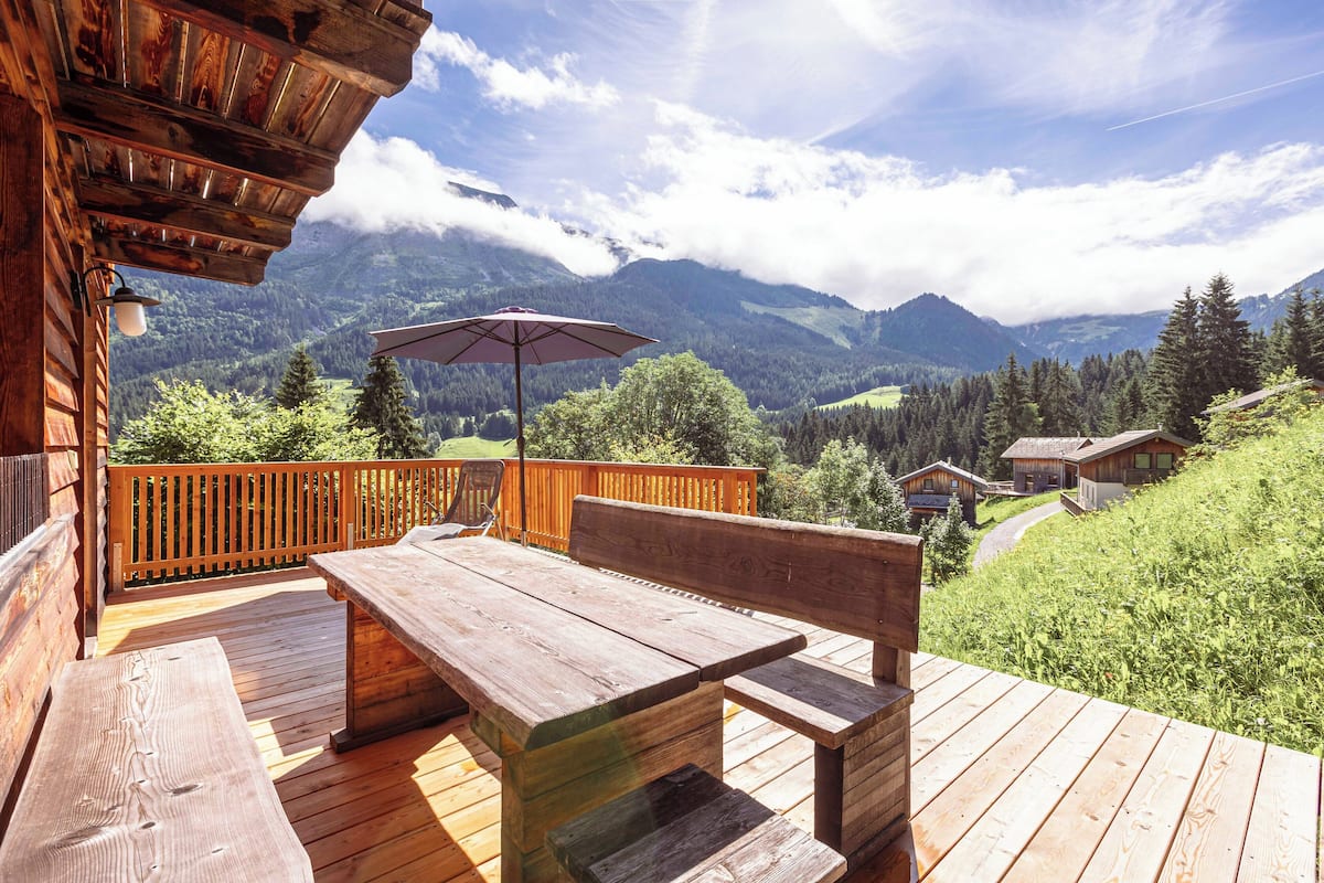 Una terraza de madera con mesa y bancos, con vistas a un paisaje montañoso.