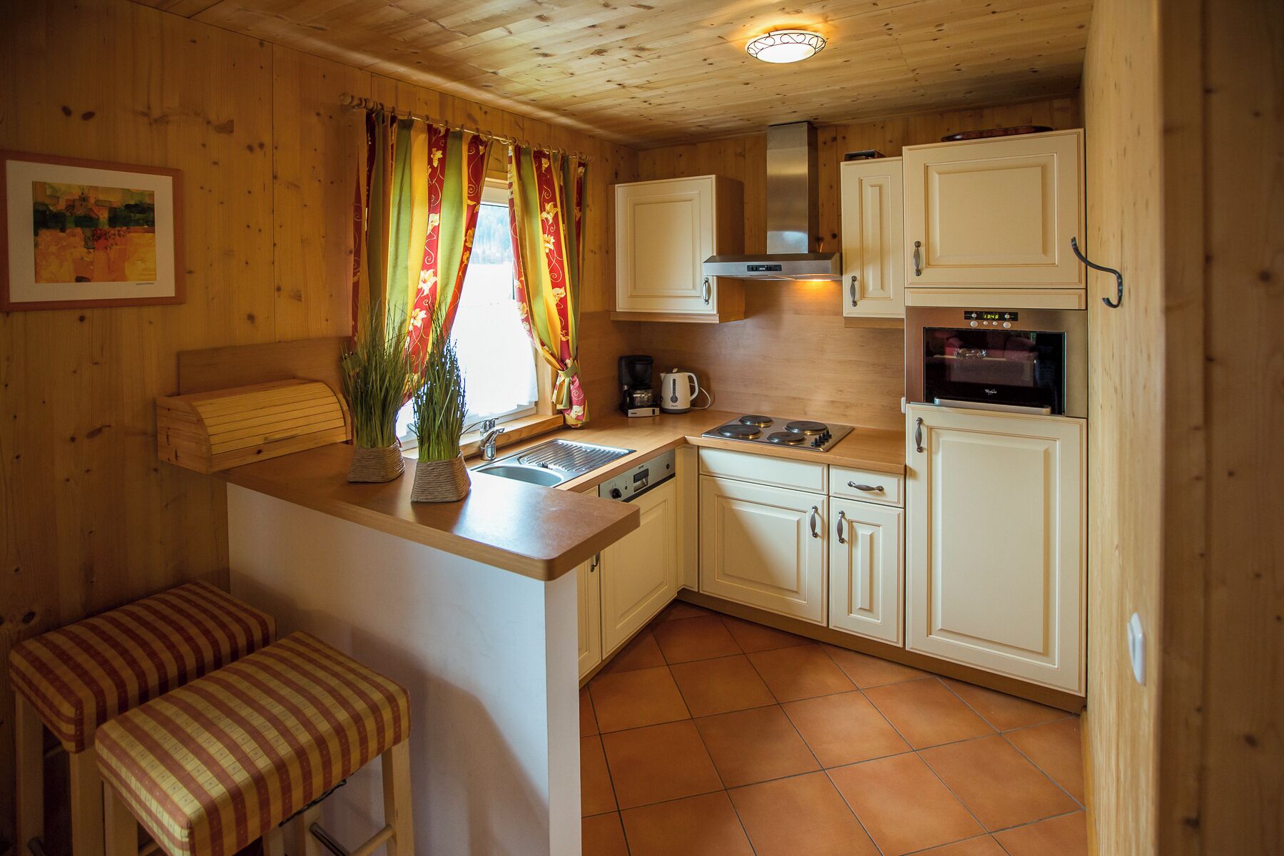 Eine kleine Küche mit Herd, Backofen und Spüle. Ein Sitzplatz und ein Fenster mit Vorhängen sind ebenfalls vorhanden.