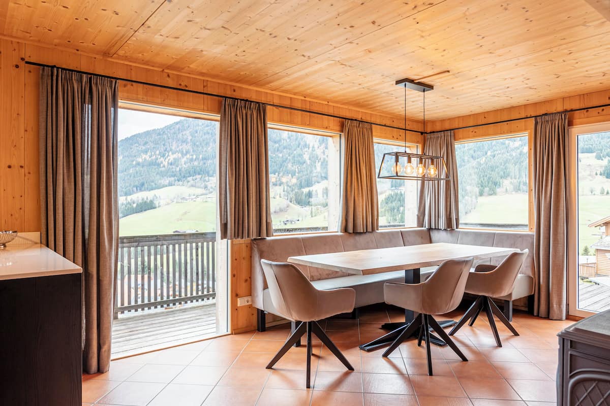 Un interior de madera con una mesa de comedor, sillas y ventanas amplias con vistas a un paisaje montañoso.