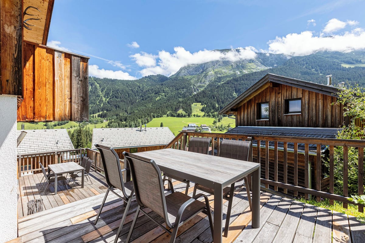 Terraza de madera con mesa y sillas, con vistas a un paisaje montañoso.