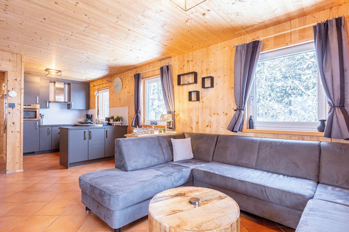Un interior de cabaña rústica moderna con un sofá seccional gris, piso de madera, una zona de cocina y ventanas amplias con cortinas.