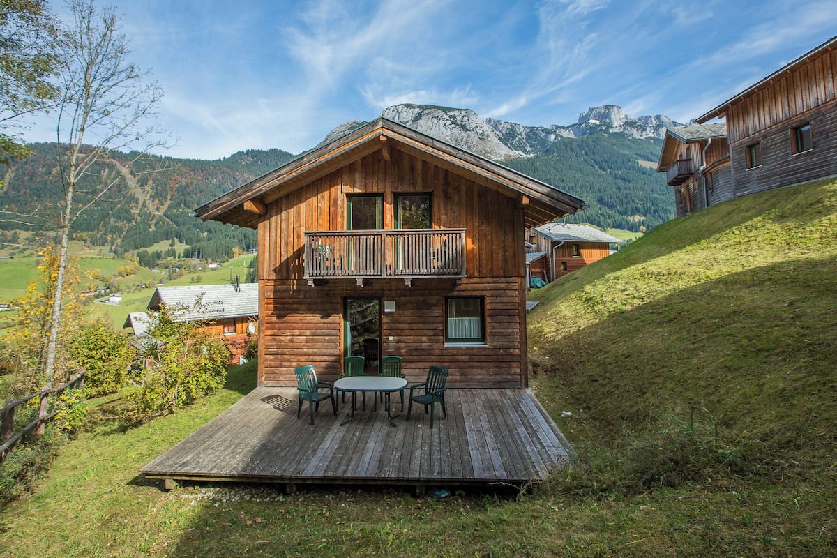 Una cabaña de madera con balcón y terraza con sillas, con montañas al fondo.