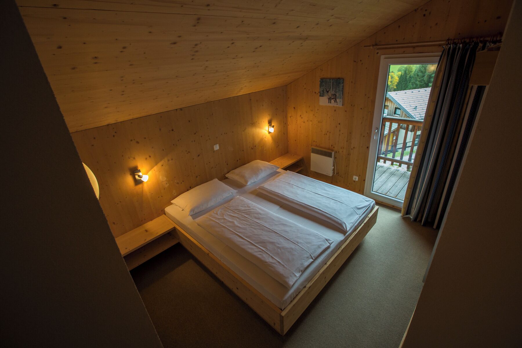 Ein Schlafzimmer einer Holzhütte mit einem Bett, zwei Wandleuchten, einem kleinen Fenster und Blick auf die Terrasse.