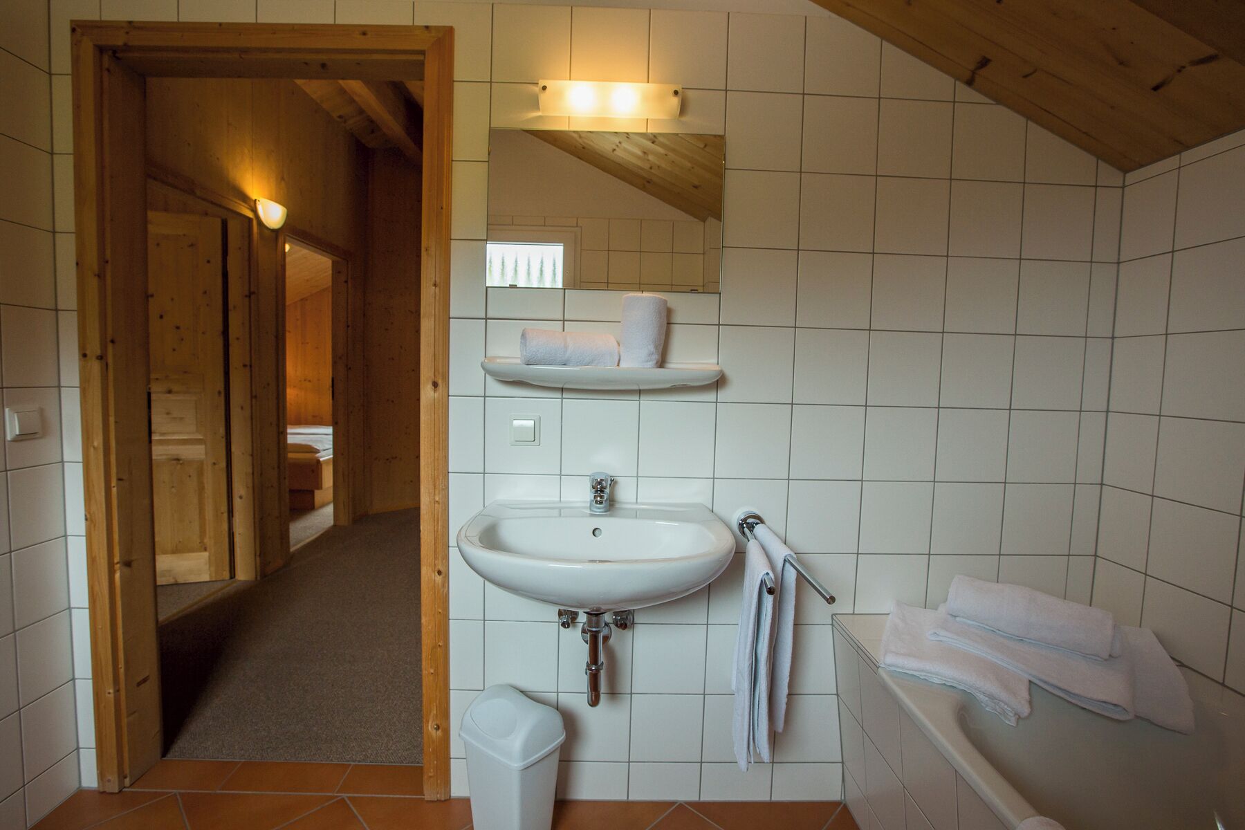 Un baño con lavamanos, inodoro y bañera, paredes de madera y un pasillo que conduce a otra habitación.