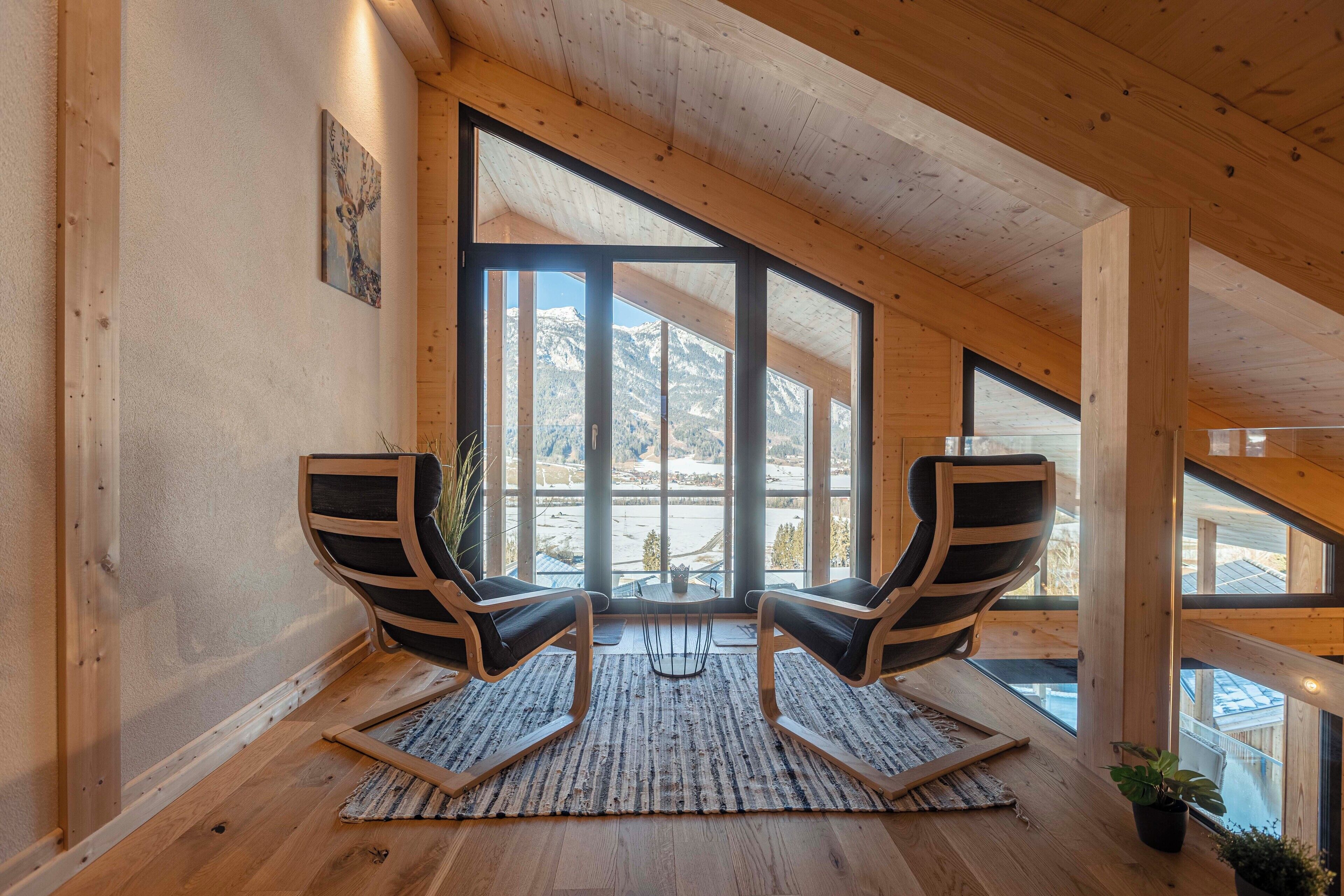 Een kamer met houten balken, grote ramen, twee zwarte leren fauteuils, een klein tafeltje en uitzicht op bergen.