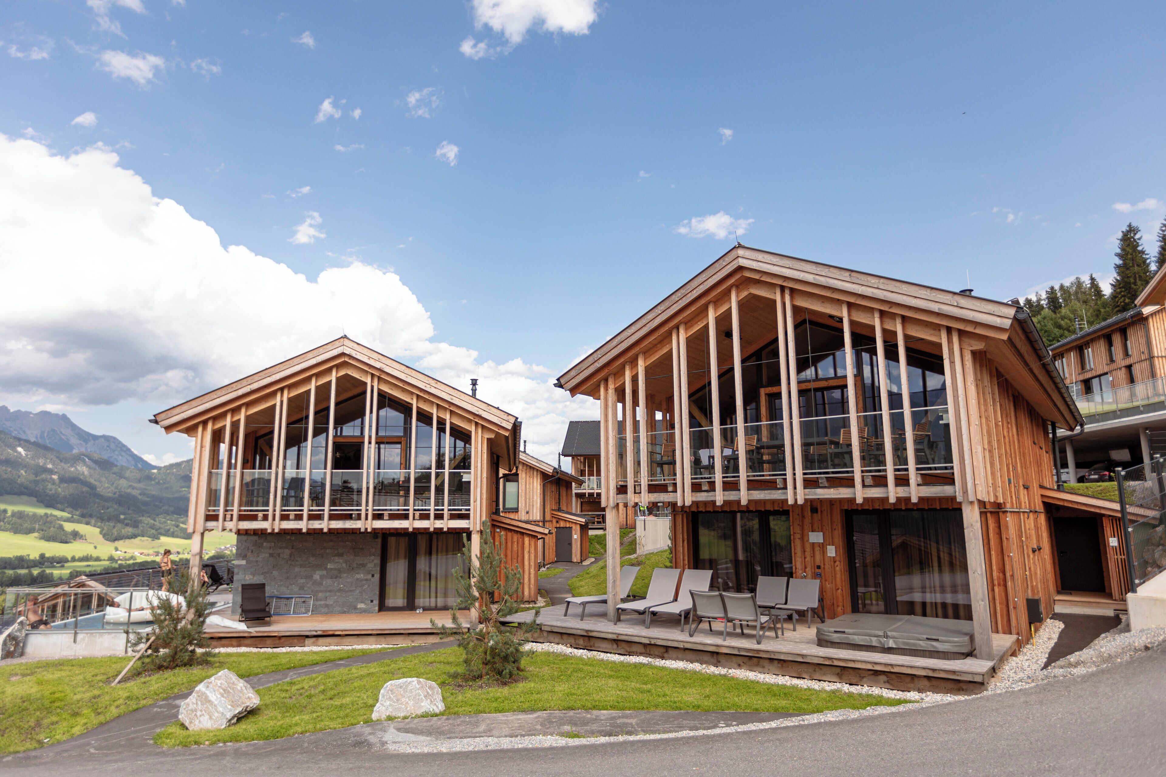 A modern wooden building with large windows, a patio with seating, and a view of the mountains.