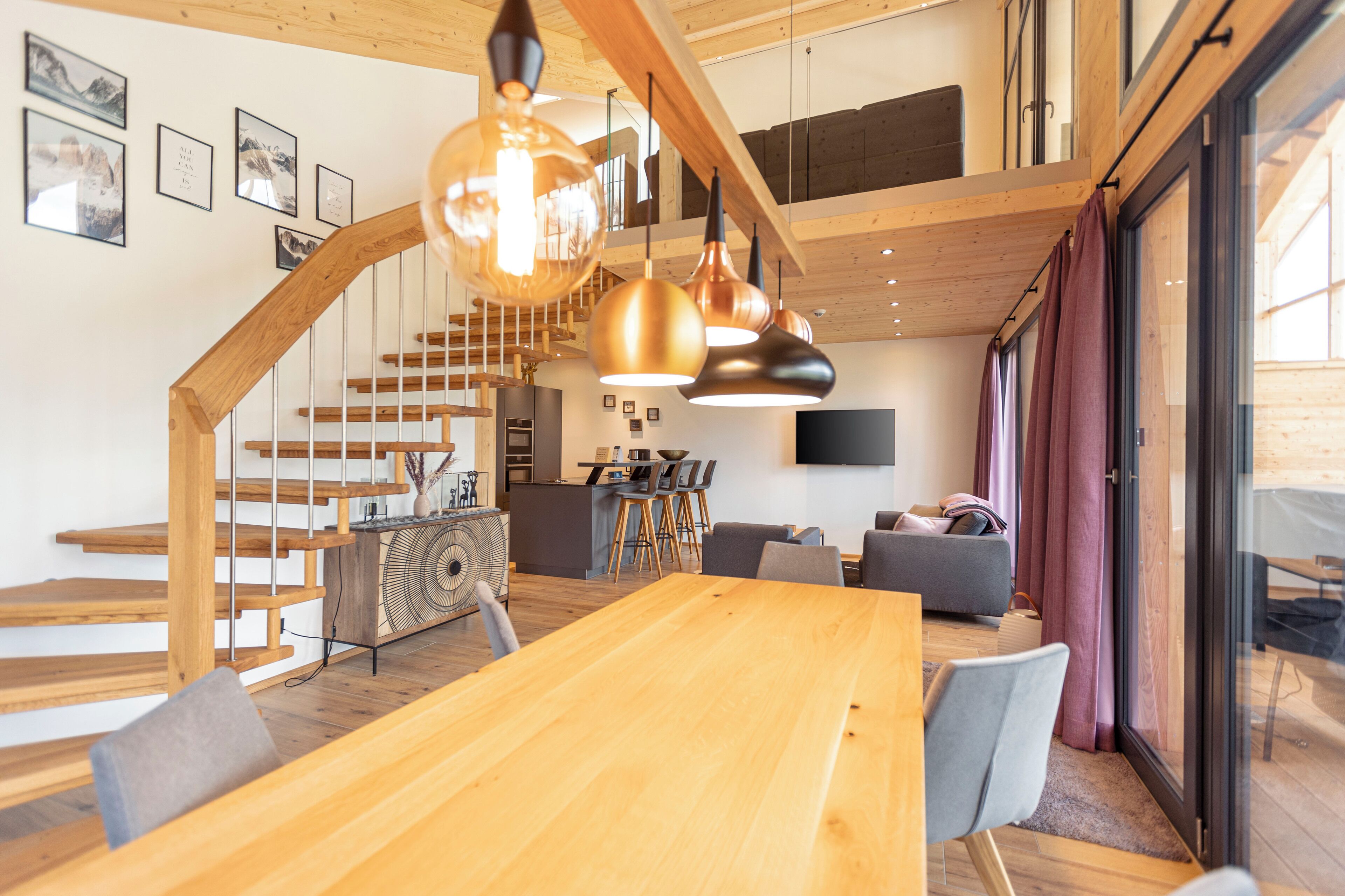 A spacious living area with a wooden staircase, a dining table, and modern pendant lighting.