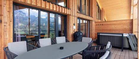 A wooden deck with a round table and chairs, large windows, and a view of mountains.