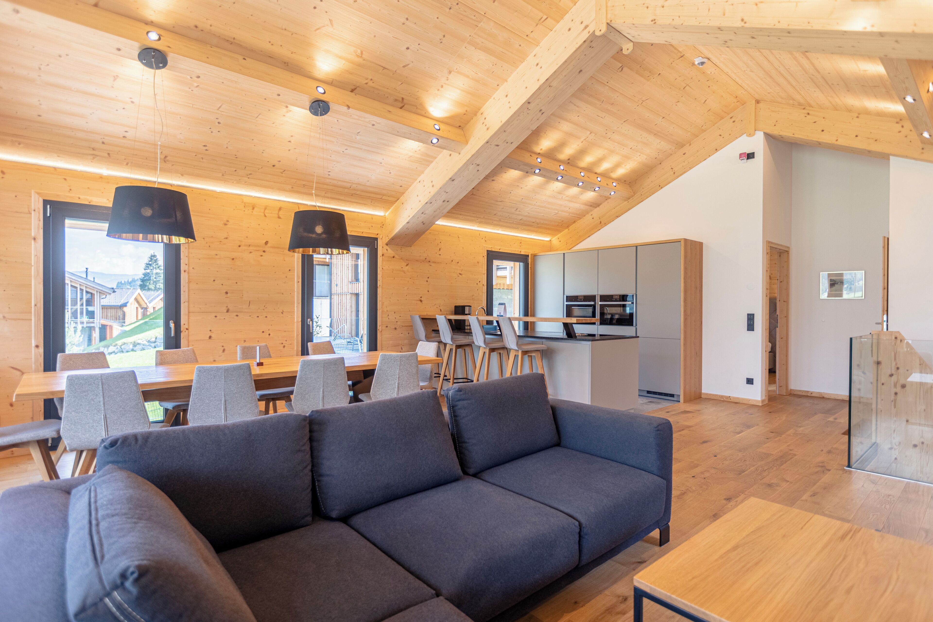 A modern interior with a wooden ceiling, a dining area with a long table and chairs, a sofa, and a kitchen area.
