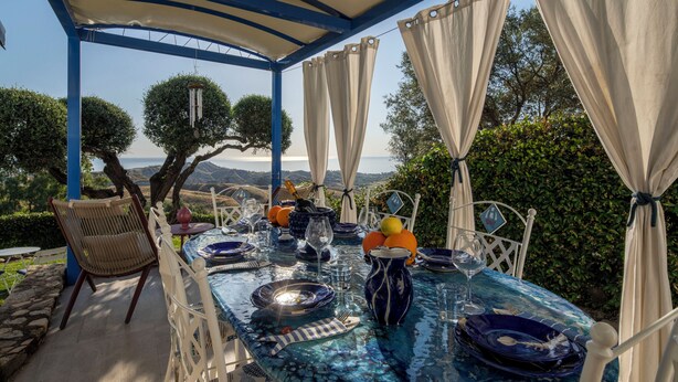 An outdoor dining area with a table set for two, surrounded by white chairs, a blue tablecloth, and a view of the landscape. - Villa Acquaro Santa Caterina  (Santa Caterina dello Ionio)