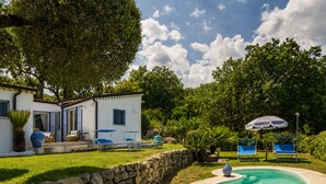 Une piscine entourée de chaises longues, un mur en pierre et un bâtiment blanc aux volets bleus.