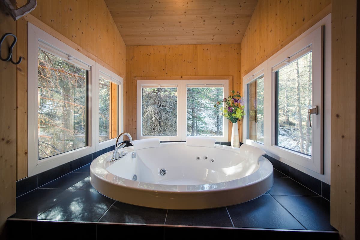 A wooden cabin with a large circular bathtub, large windows, and a view of the forest.
