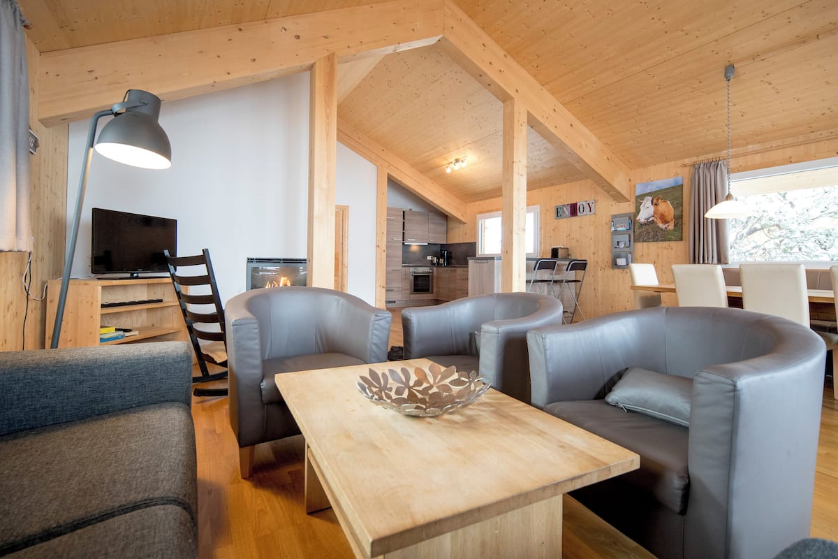 A cozy cabin interior with a dining area, two sofas, a wooden table, and a kitchen in the background.