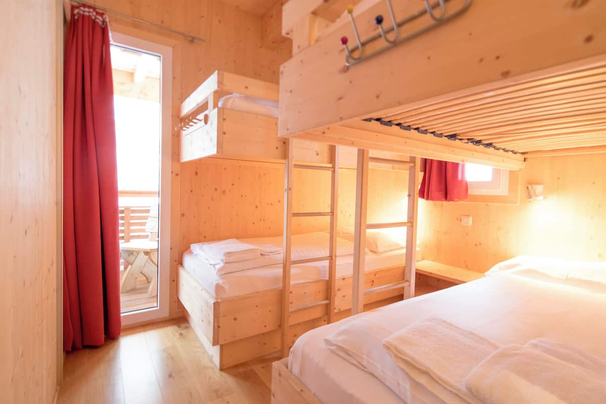 A wooden cabin with bunk beds, a window with red curtains, and a wooden table outside.