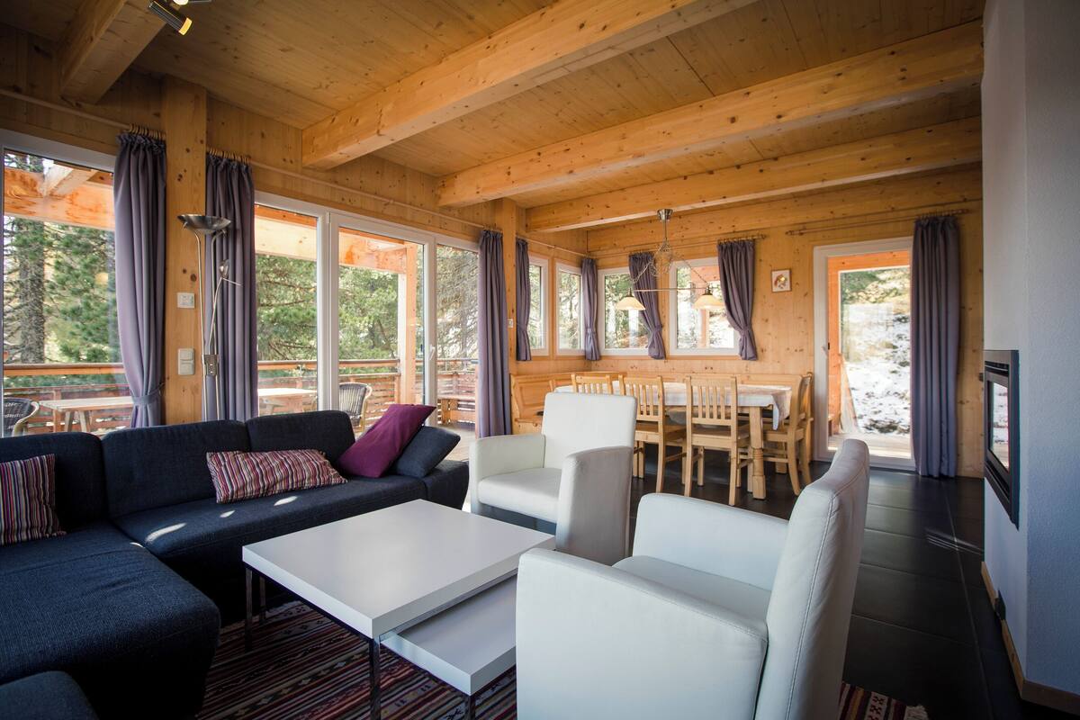 A cozy cabin interior with a sofa, chairs, a dining table, and large windows.