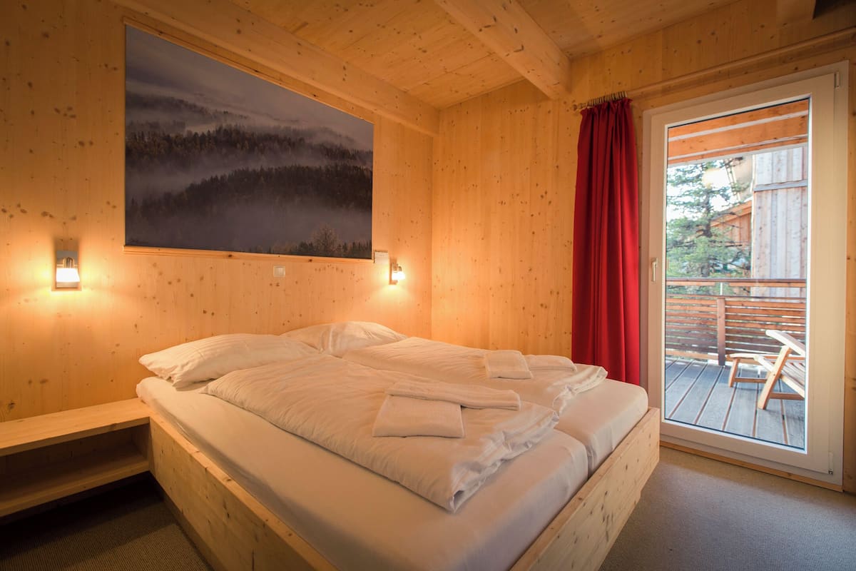 A wooden room with a bed, a painting of a forest, and a view of a deck outside.