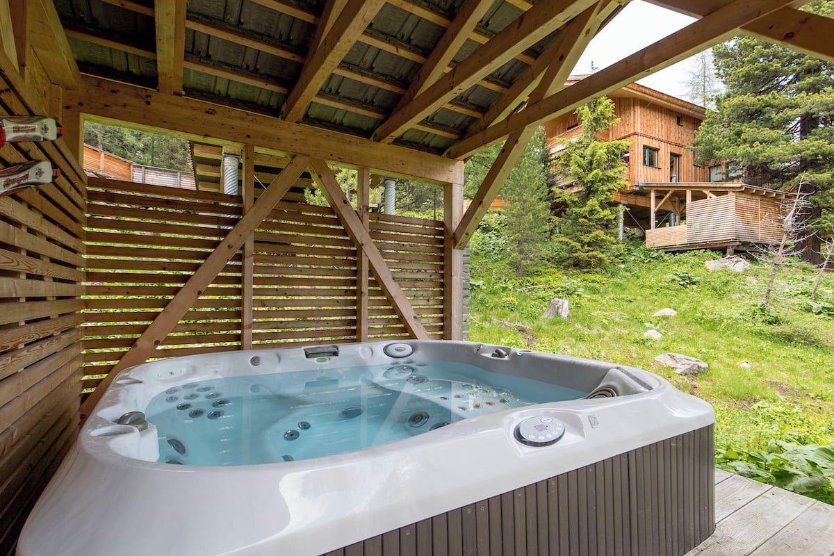 A wooden deck with a hot tub, surrounded by lush greenery and a wooden cabin in the background.