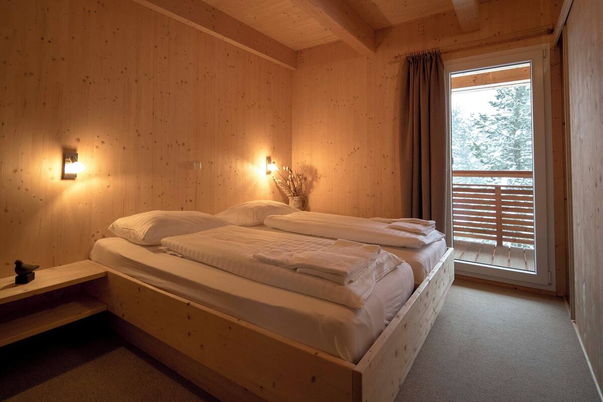 A wooden cabin bedroom with a bed, bedside lamps, and a window with blinds.